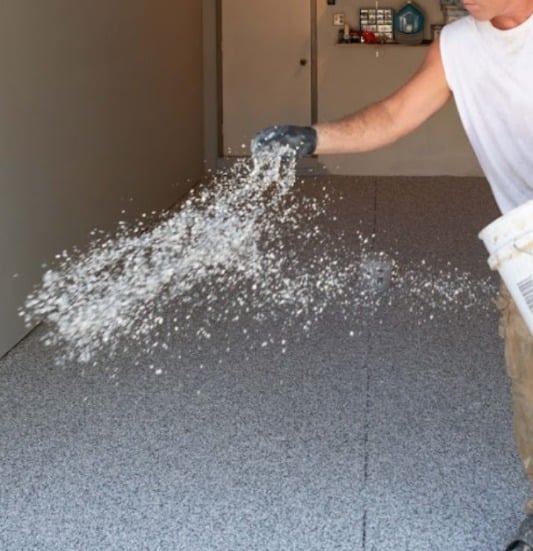A person is scattering small white pellets across a gray speckled floor, creating a dynamic arc of particles mid-air, likely in a garage.