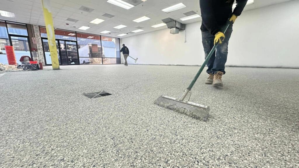 Two people in a large room with a speckled floor are using long-handled squeegees. Construction materials are visible near large windows in the background.