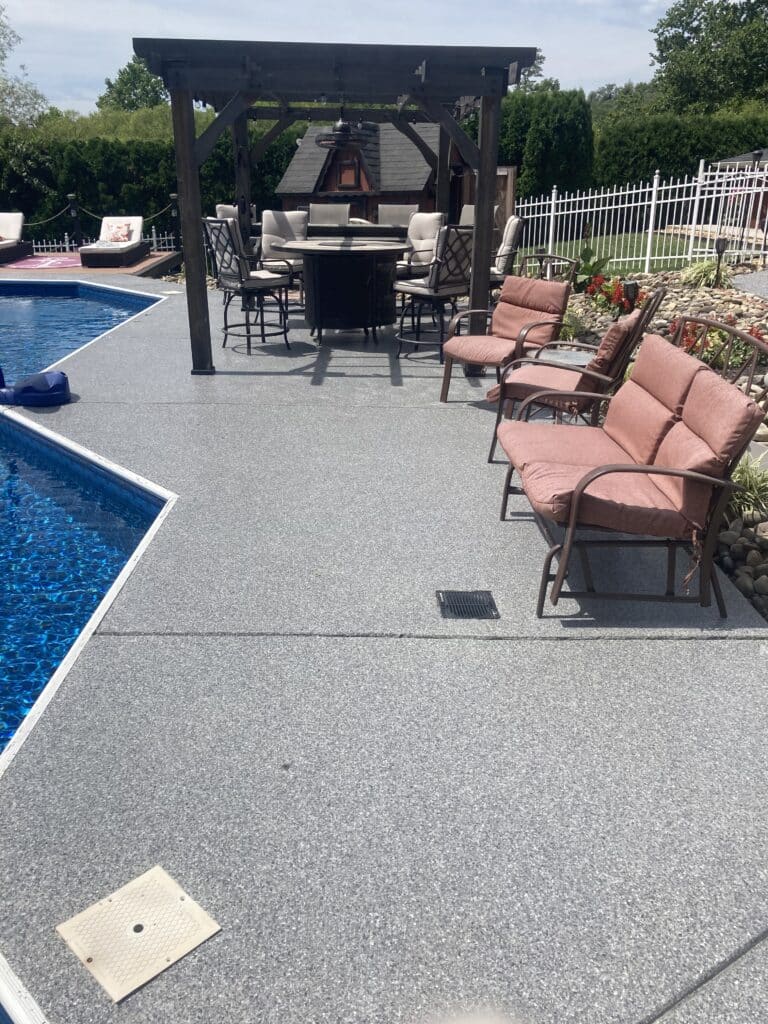 An outdoor area with a pool and a pergola housing a dining set. Pink cushioned chairs line the textured patio. A white fence surrounds the space.