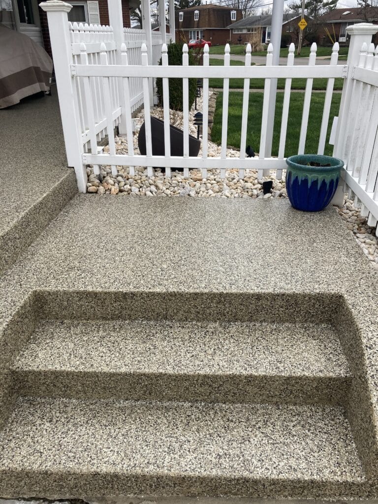 A textured outdoor staircase leads down to a walkway enclosed by a white picket fence. A blue planter and landscaping are visible on the sides.