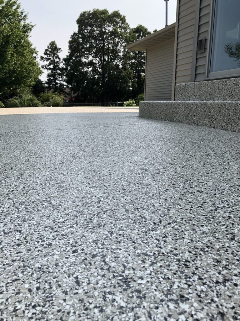 This image shows a speckled epoxy-coated floor extending to a building with beige sidings and a white framed window, with trees visible in the background.