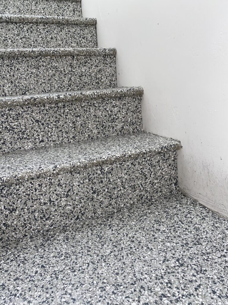 A close-up of a flight of outdoor terrazzo stairs with grey and white speckled steps against a white wall, leading upwards to the left.