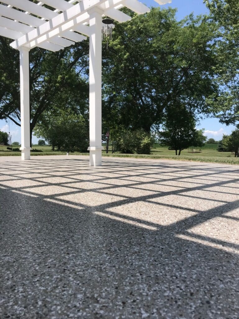 This image shows a ground-level view of a white pergola casting geometric shadows on a textured ground, with lush green trees and a clear sky in the background.