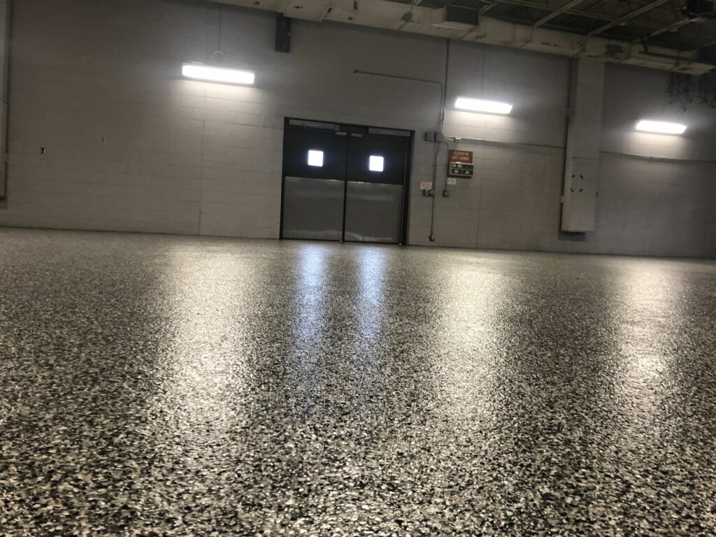 This image shows an empty industrial space with shiny speckled flooring, white walls, fluorescent lights, and a pair of windows on a door.