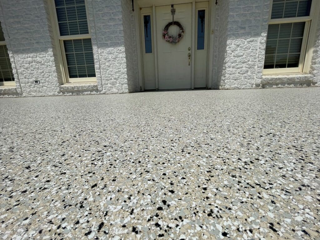 The image shows an exterior view of a building with a white door, flanked by windows, a wreath hanging, and a textured driveway surface in the foreground.