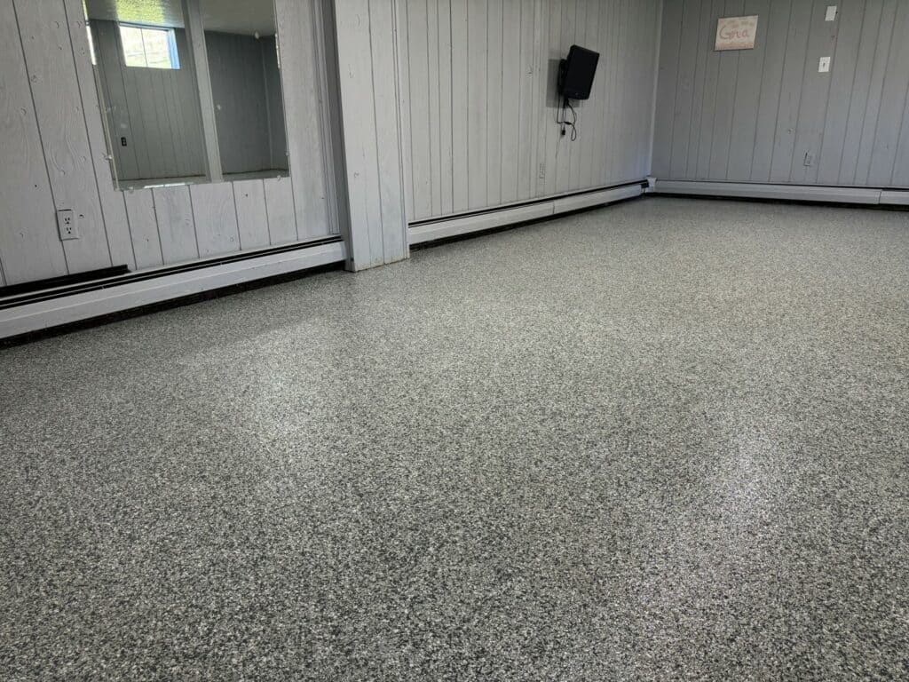 An empty room with gray speckled flooring, pale wood walls, baseboard heaters, a mirror, a wall-mounted TV, and a sign with cursive text.